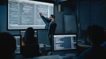 Professor stands in front of a class gesturing to a projector screen full of code to deliver a software lecture.